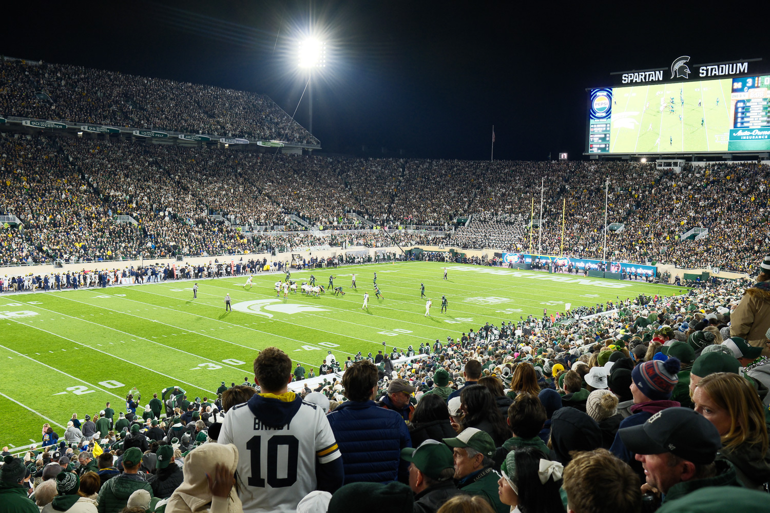 A crowded sports stadium at night, with a football game in progress on the field and spectators filling the stands.