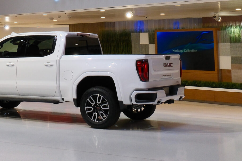 A white GMC pickup truck with large tires and alloy wheels is displayed in a showroom with a "Heritage Collection" sign in the background.