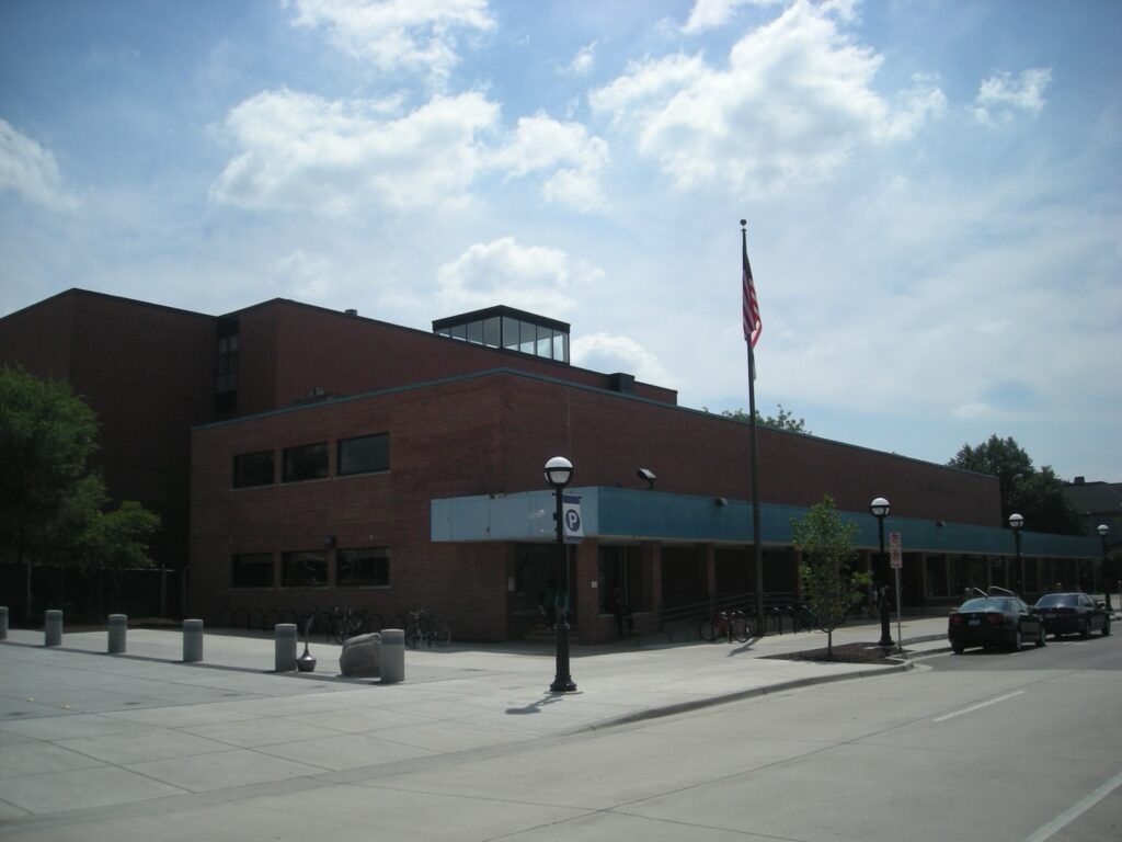 A brick building with an American flag flying, parking meters, and parked cars in front, suggesting a library or community center in Ann Arbor.