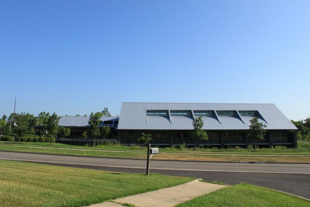 A large modern library building with a sloped metal roof, surrounded by green grass and trees, and located near a road.