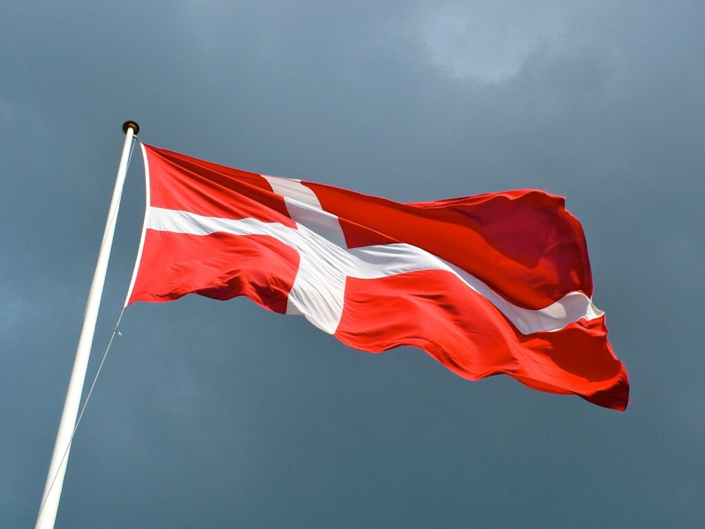 The image shows the vibrant red and white national flag of Denmark waving against a cloudy blue sky.