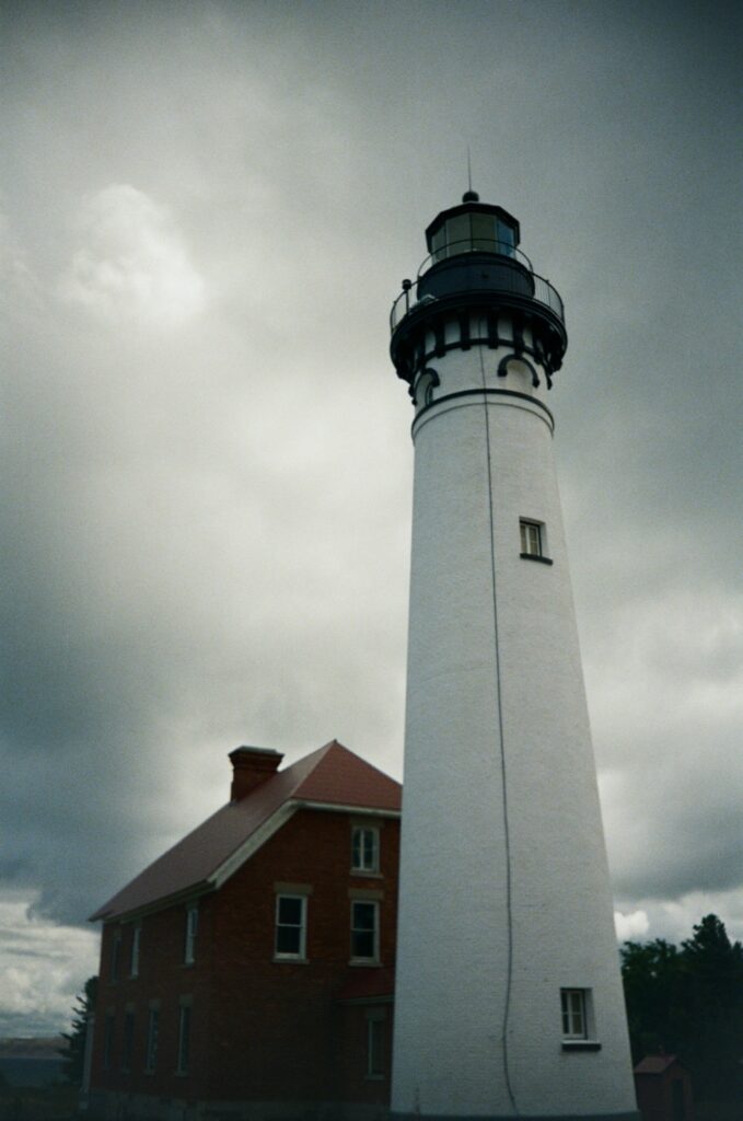 A tall, white lighthouse stands against a cloudy, overcast sky, surrounded by a building with a red roof.