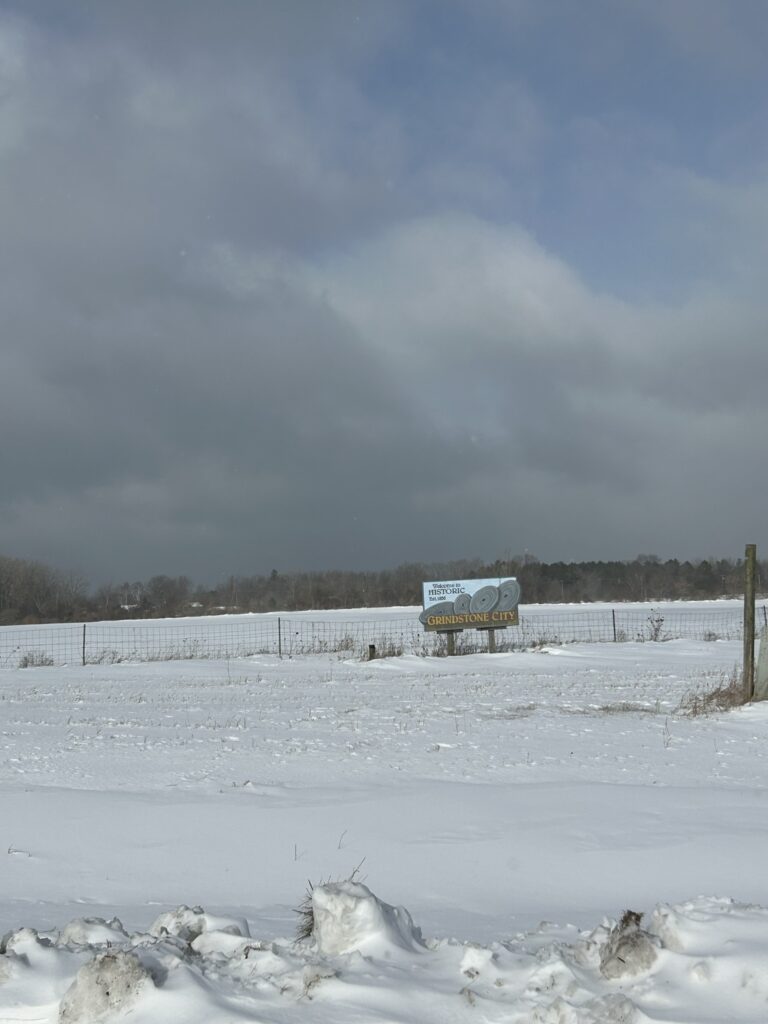 A lone sign marking "Grindstone City" stands along a snow-covered road, with the gray expanse of Lake Huron visible in the distance.