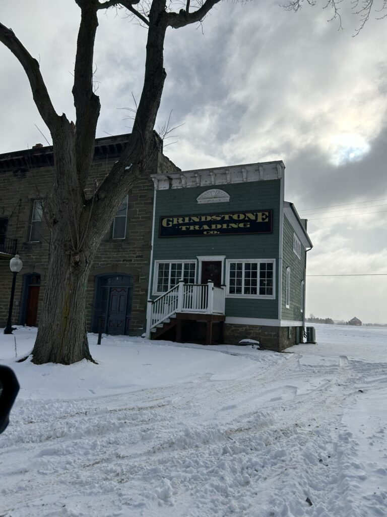 A snow-covered building with the name "Grindstone City" painted on its side stands in a wintry landscape, reflecting the town's history as a former center of grindstone production.
