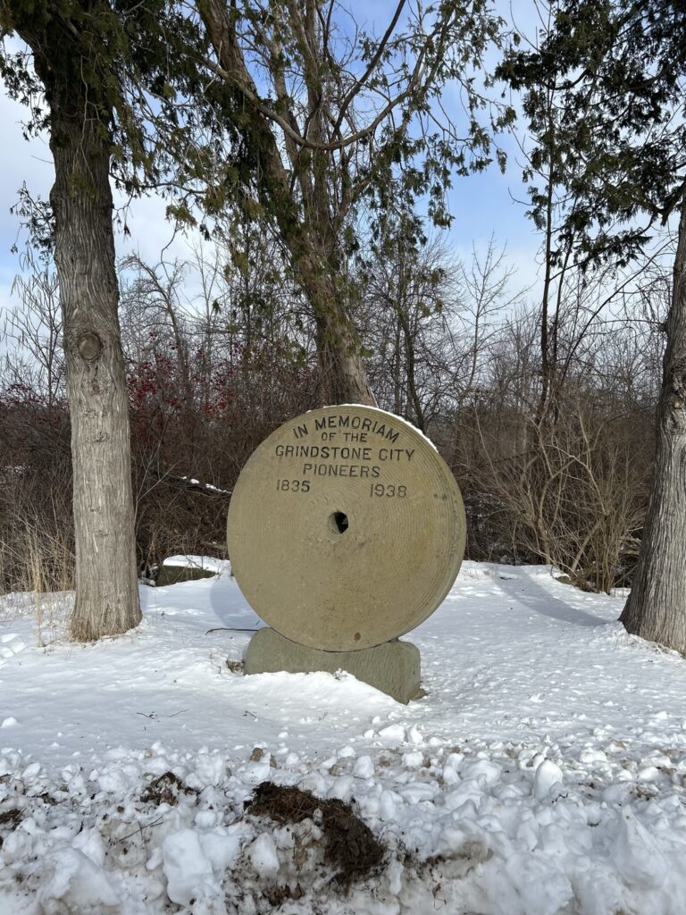 In the image, a large grindstone marker stands in the snow, with trees and a snowy landscape in the background. The marker reads "In Memoriam of the Grindstone City Pioneers 1835 1938".