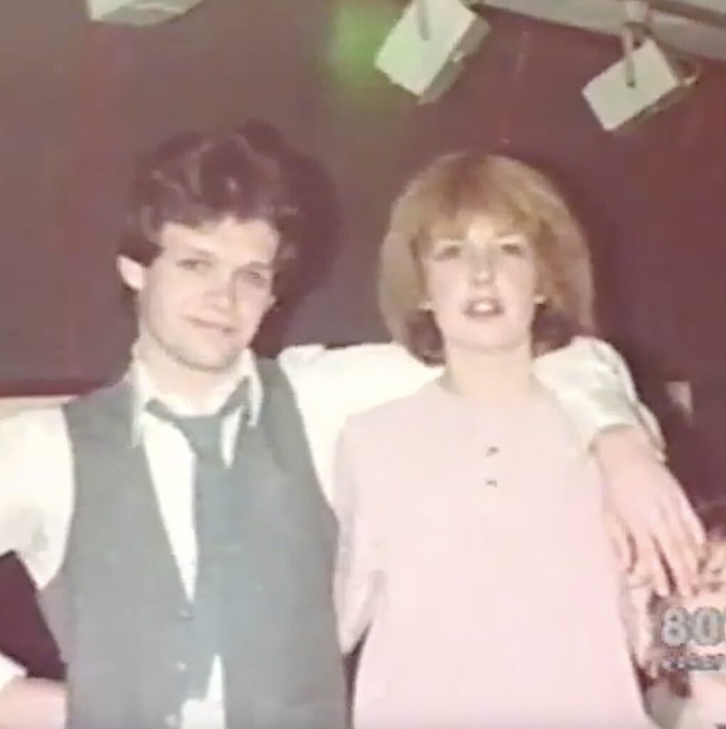 Two people, a man and a woman, appear to be standing together in a room. The man has short dark hair and is wearing a light-colored jacket, while the woman has blonde hair and is wearing a white blouse.