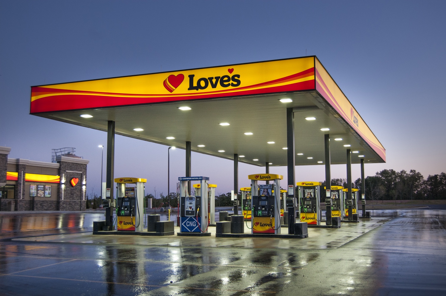 A brightly lit Loves gas station at dusk, with rows of fuel pumps and the distinctive Loves logo and branding prominently displayed.