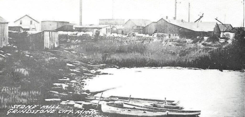 A black and white image showing the shoreline of Grindstone City, Michigan, with buildings, smokestacks, and a boat on the frozen lake. The town was once a thriving hub for grindstone quarrying and production.