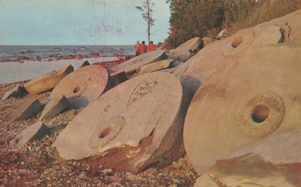 Large, weathered grindstones rest on the rocky shore, with a few people visible in the distance on the bluffs overlooking Lake Huron.