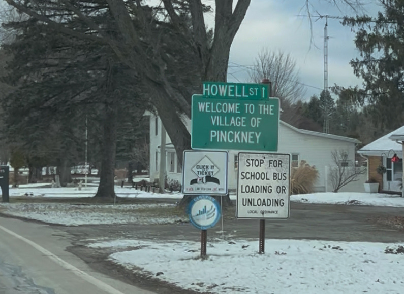 A snowy street scene with a "Welcome to the Village of Pinckney" sign, a "Stop for school bus" sign, and a tree-lined road in the background.
