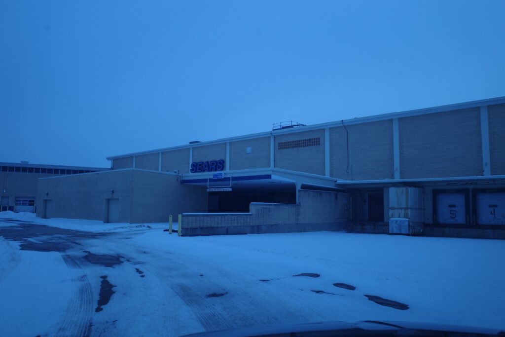 Exterior of a large, abandoned Sears retail store in Livonia, Michigan, with a snowy parking lot in the foreground.