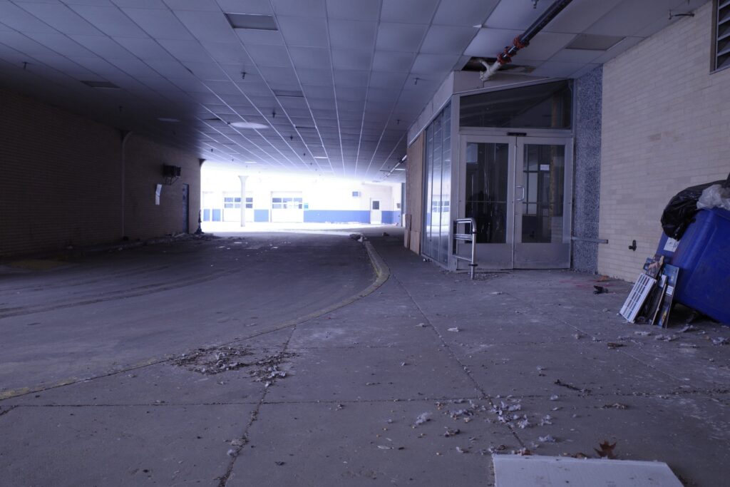 A long, dimly lit hallway with peeling walls and debris strewn across the concrete floor, leading to a glass-paneled entryway in a dilapidated building.