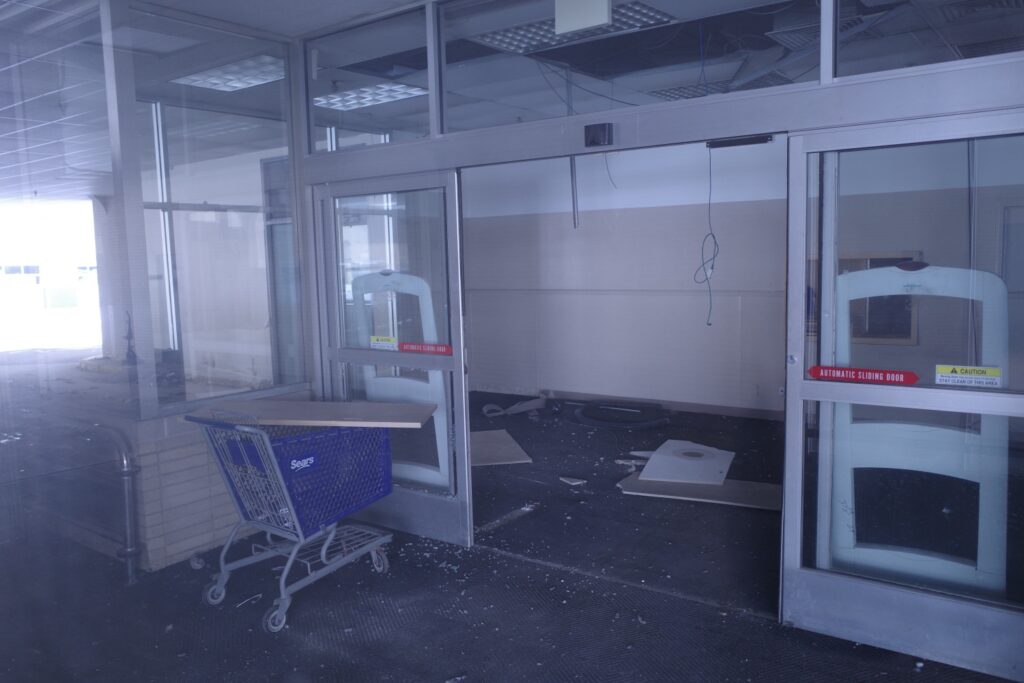 A deserted Sears store interior with a shopping cart and shattered glass on the floor, suggesting abandonment.