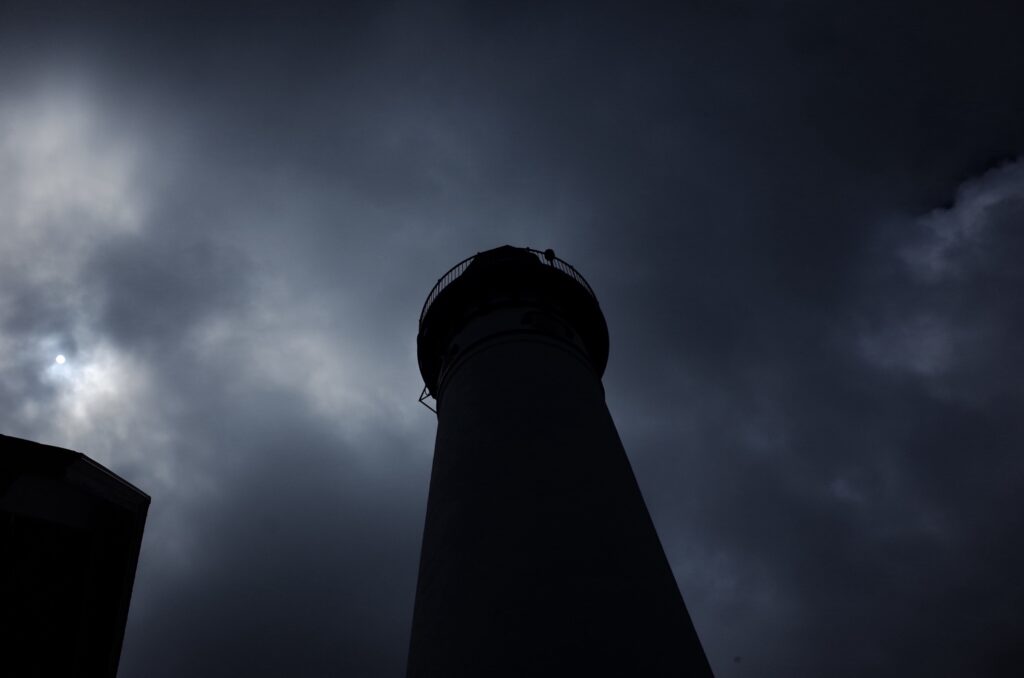A dark, ominous-looking lighthouse tower stands against a stormy sky with clouds.