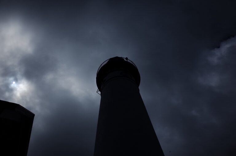 A dark, ominous-looking lighthouse tower stands against a stormy sky with clouds.