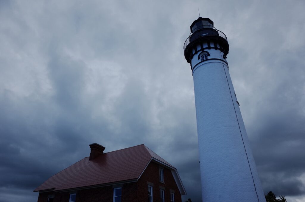 A tall, blue lighthouse stands against a cloudy, stormy sky, with a nearby building with a red roof.