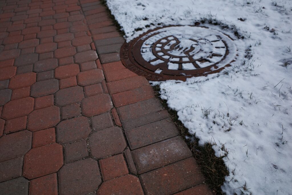 A snow-covered brick walkway with a manhole cover or utility access point in the foreground, indicating the use of heated sidewalks in Holland, Michigan.