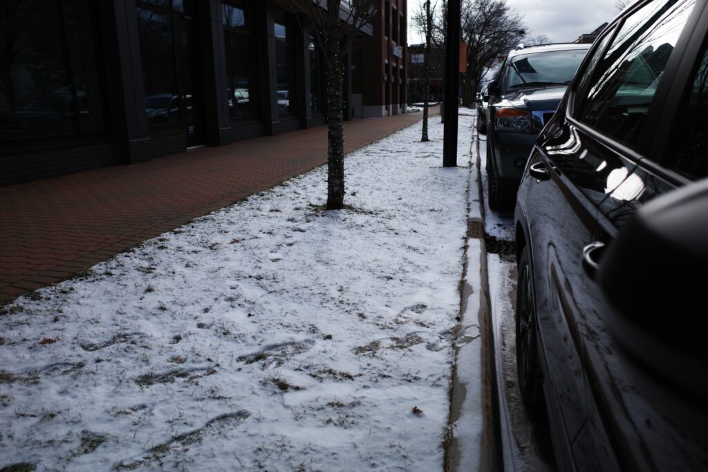 A snowy sidewalk in a town, with trees lining the street and parked cars along the curb.