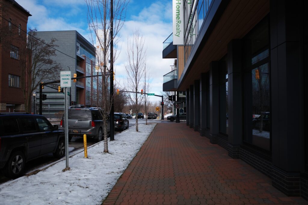 A snowy street in Holland, Michigan with brick sidewalks and heated to melt the snow, surrounded by buildings and trees.