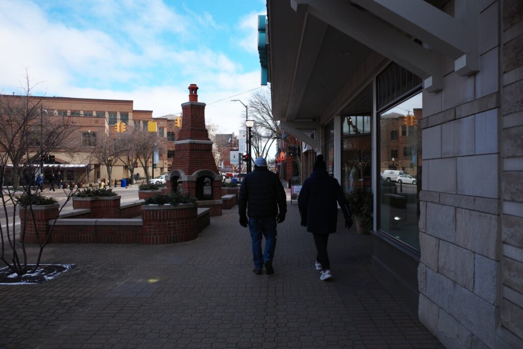 Two people walking on a brick-paved sidewalk with a large brick chimney-like structure in the background, in a downtown area with storefronts.