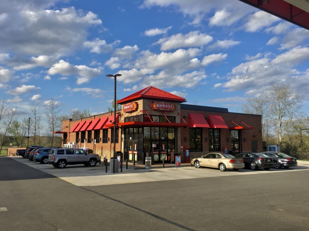 A brick building with a red roof and awning, displaying the Sheetz logo. Several cars are parked in the lot in front of the building.