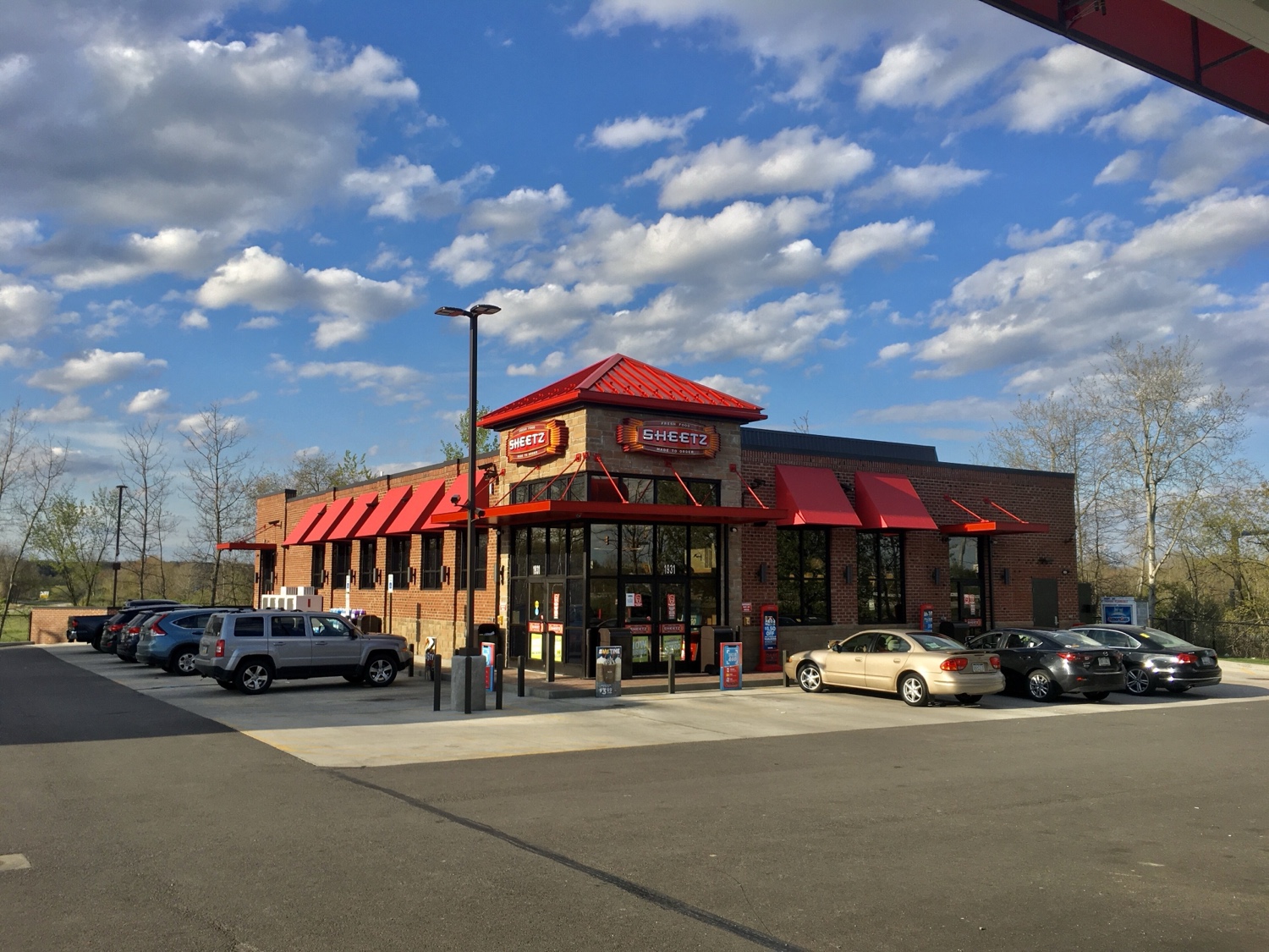 A brick building with a red roof and awning, displaying the Sheetz logo. Several cars are parked in the lot in front of the building.