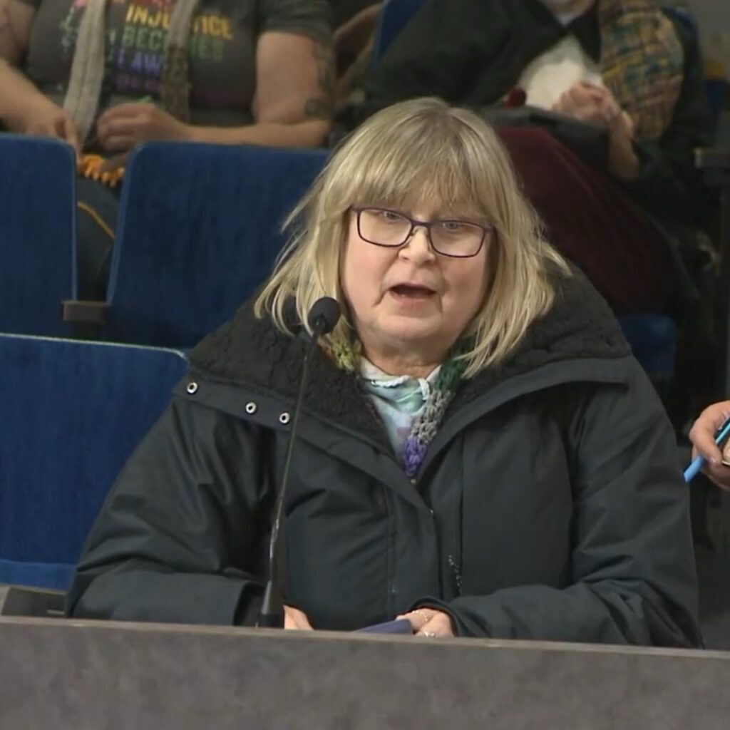 An older woman with glasses and blonde hair standing at a podium and speaking at a city council meeting.