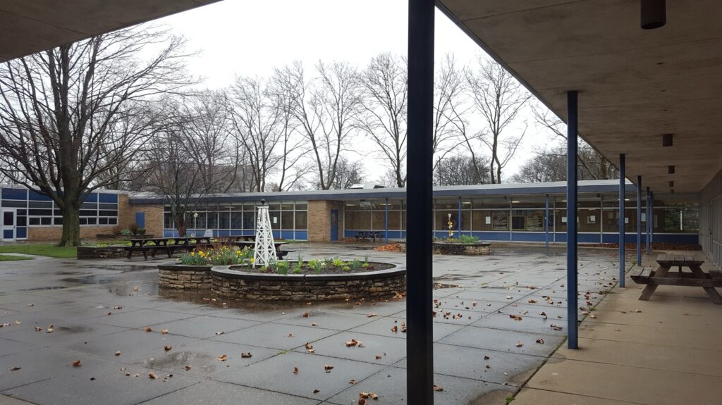 Outdoor view of a school building with bare trees and a stone planter with flowers in the foreground.