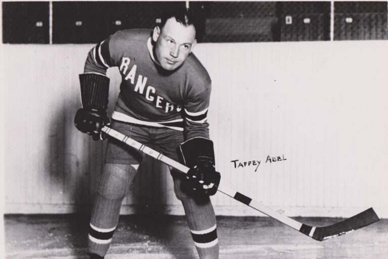 A black and white photo of a hockey player in uniform, identified as Taffy Abel, the first Native American to carry the U.S. flag at the 1924 Winter Olympics.