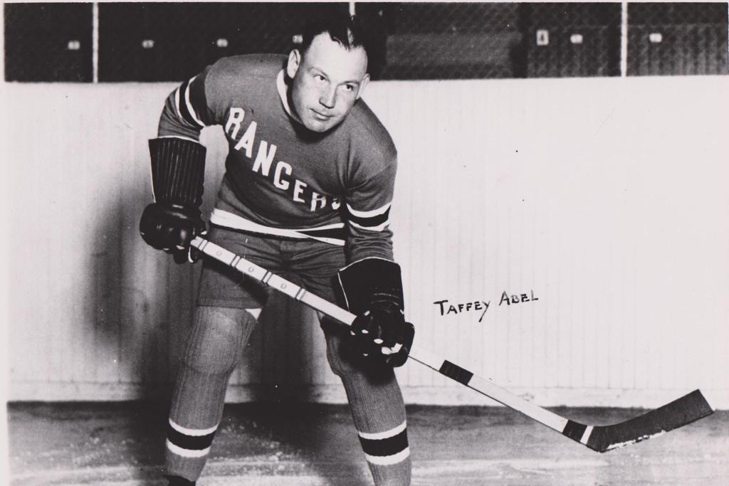 A black and white photo of a hockey player in uniform, identified as Taffy Abel, the first Native American to carry the U.S. flag at the 1924 Winter Olympics.