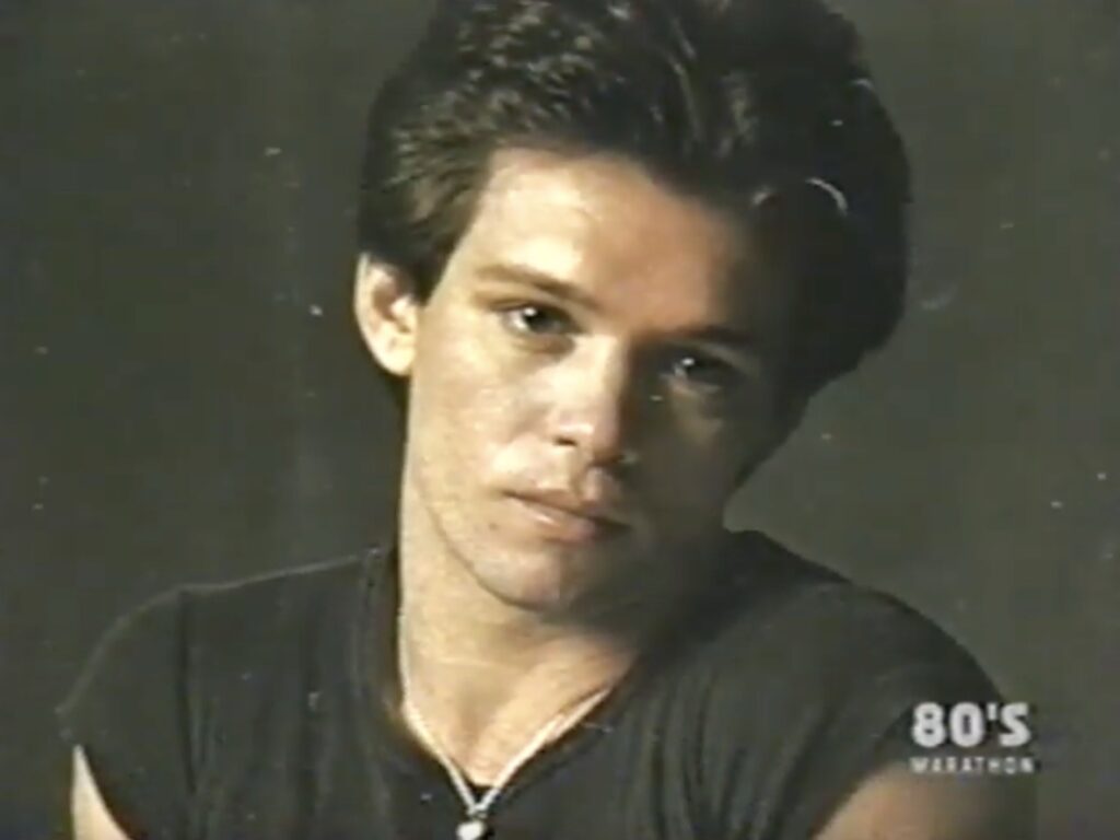 The image shows a portrait of a man with short dark hair, wearing a dark jacket and looking directly at the camera with a serious expression.