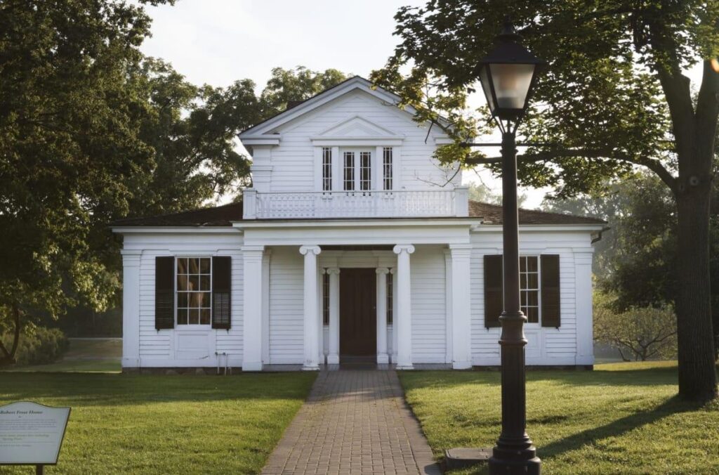 A historic white wooden house with a covered porch, surrounded by trees on a grassy lawn, with a lamp post in the foreground. This is the Washtenaw Street house where poet Robert Frost resided during his residencies at the University of Michigan in the 1920s.