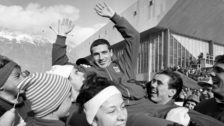 This black and white image shows a crowd of people enthusiastically greeting and celebrating a smiling man wearing an Olympic athletic uniform, the American speed skater Terry McDermott who won gold at the 1964 Winter Olympics.